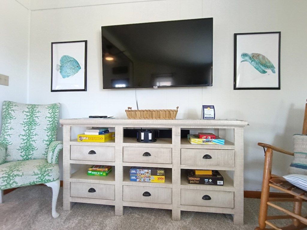 Una sala de estar con un televisor montado en la pared, un mueble de madera con cajones, un sillón estampado y un cuadro enmarcado de una tortuga.