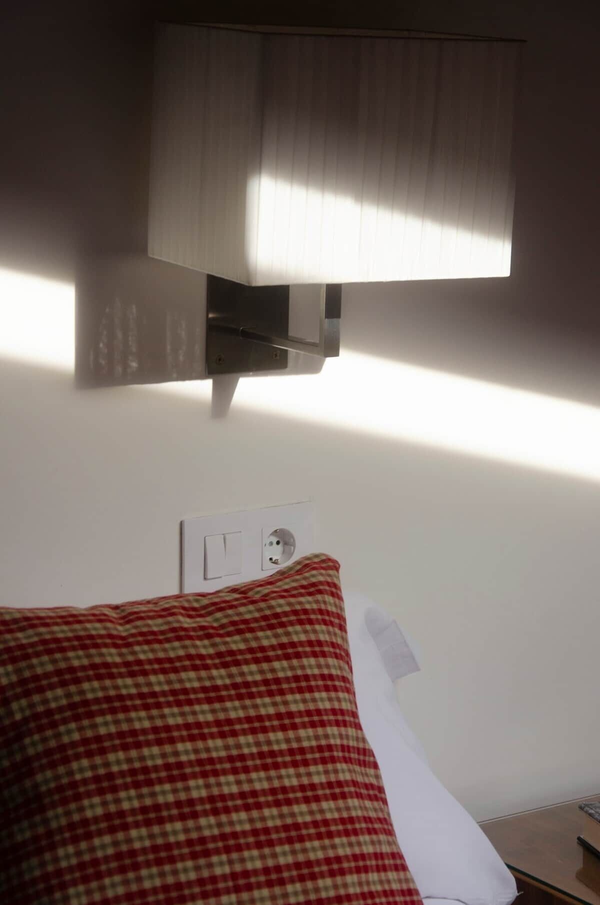Une chambre d’hôtel avec un lit, une lampe, une prise de courant et une fenêtre avec des stores.