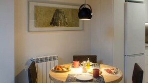 A dining area with a round table, chairs, a pendant light, a framed picture, and a refrigerator in the background.