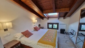 A bedroom with a wooden ceiling, a bed with pillows, a bedside table, a lamp, a radiator, and a window.