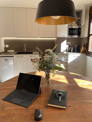 A modern kitchen with a laptop, a mouse, and a notebook on a wooden table, a vase with flowers, and a pendant light.