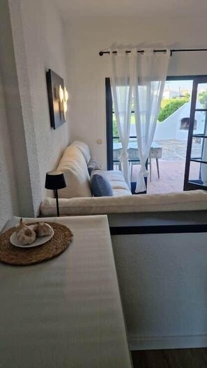 Ein Zimmer mit einer Couch, einem kleinen Tisch mit Unterlage, einer Lampe und Blick durch ein Fenster mit Vorhängen.