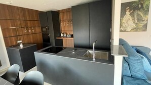 Et moderne køkken med mørke skabe, en central ø med vask, og en siddeplads med en blå sofa.