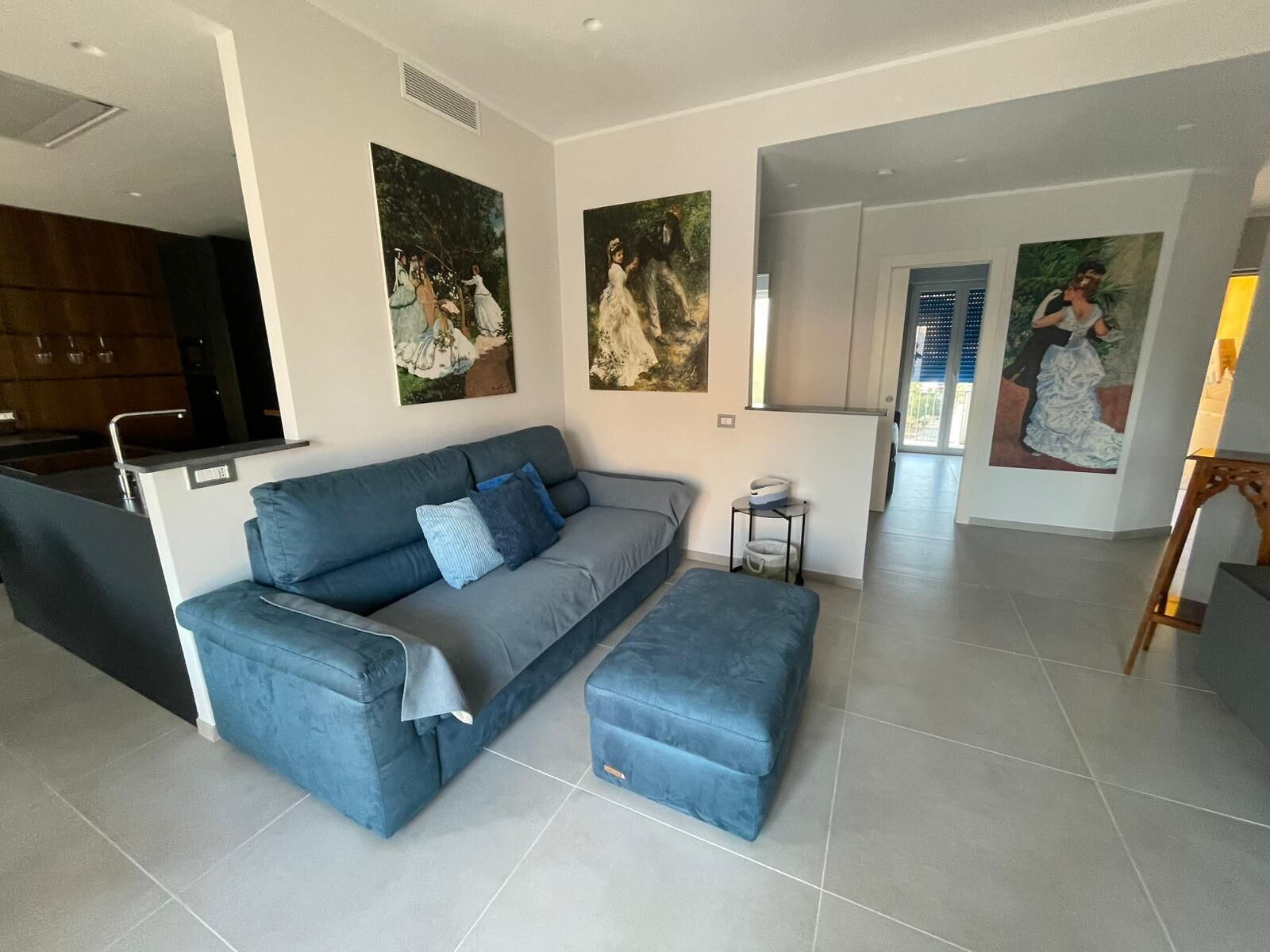 A modern living room with a grey sofa, a matching ottoman, and a dining area with a kitchen in the background.