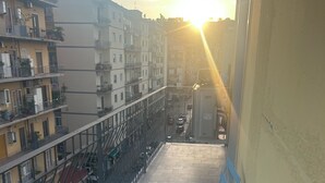 En balkon med et rundt bord og blå stole, med udsigt over en gade med parkerede biler og bygninger.