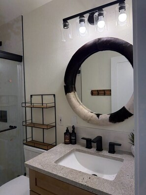 A bathroom with a large round mirror, a sink, a shelf with toiletries, and a glass shower enclosure.
