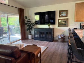 A living room with a TV, a dining area with bar stools, a sofa, and a large window.
