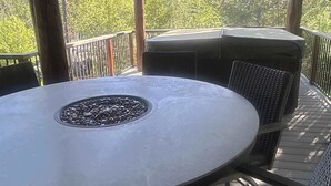 En overdækket terrasse med et rundt bord og stole, en bålplads og udsigt til træer.