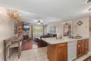 A modern kitchen with a central island, a dining area with a wooden table and benches, a living room with a sofa and fireplace, and a ceiling fan. - Mountain View Memories Unit #2102 (Gatlinburg)