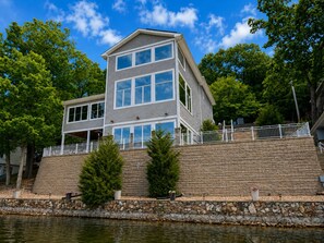 A multi-story house with a stone retaining wall, a balcony, and a view of a body of water. - Lake Paradise Lakefront Stay (Eldon)