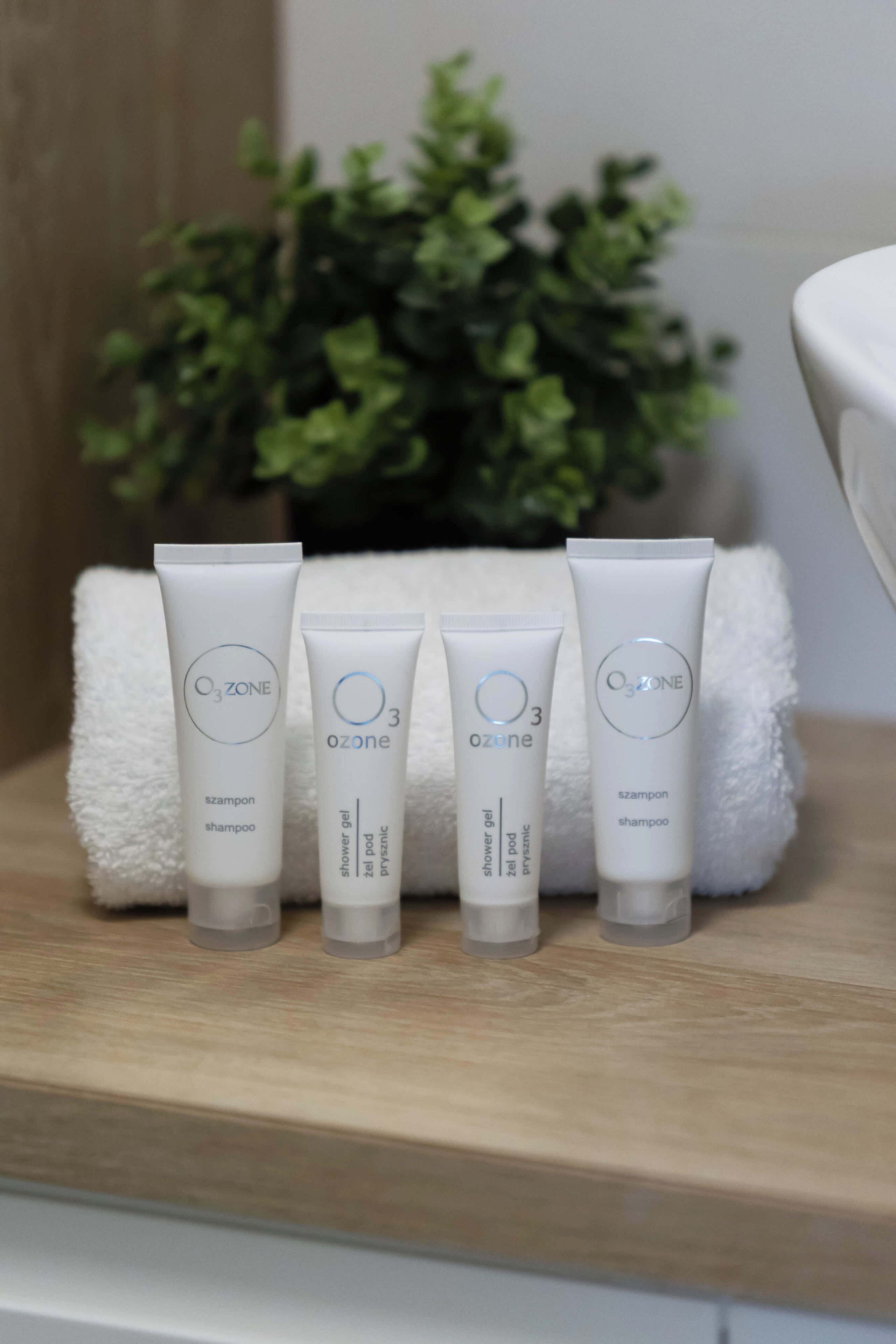 Quatre produits de toilette de la marque OZONE sont disposés sur une surface en bois, avec une serviette blanche et une plante verte en arrière-plan.