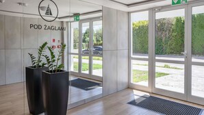 Een moderne ingang met een bord waarop 'POD ŽAGLAMI' staat en een logo erboven. Er staan twee grote plantenbakken met groene planten, een rookmelder en een groen nooduitgangsbord.