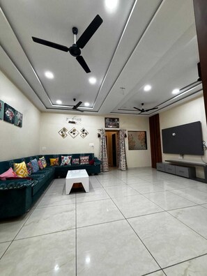 Una sala de estar con un sofá modular, una mesa de centro, ventiladores de techo, iluminación empotrada, un televisor de pantalla plana y un mueble de televisión montado en la pared.