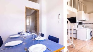 Un coin repas avec une table bleue dressée pour six personnes, une cuisine équipée d’un lave-linge et un escalier menant à l’étage supérieur.