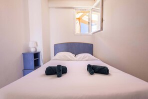 A hotel room with a bed, two rolled towels, a nightstand, a lamp, and a window with a view. - The Holiday Corner (Valras-Plage)