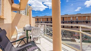 Un balcon avec une table et des chaises, une vue sur un immeuble situé de l’autre côté de la rue, et un ciel bleu dégagé parsemé de nuages.