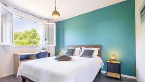 A bedroom with a bed, a desk, a nightstand, and a window with a view of greenery.