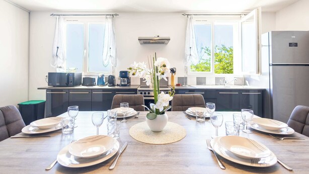 A dining area with a table set for six, a kitchen in the background, and a view of greenery outside.