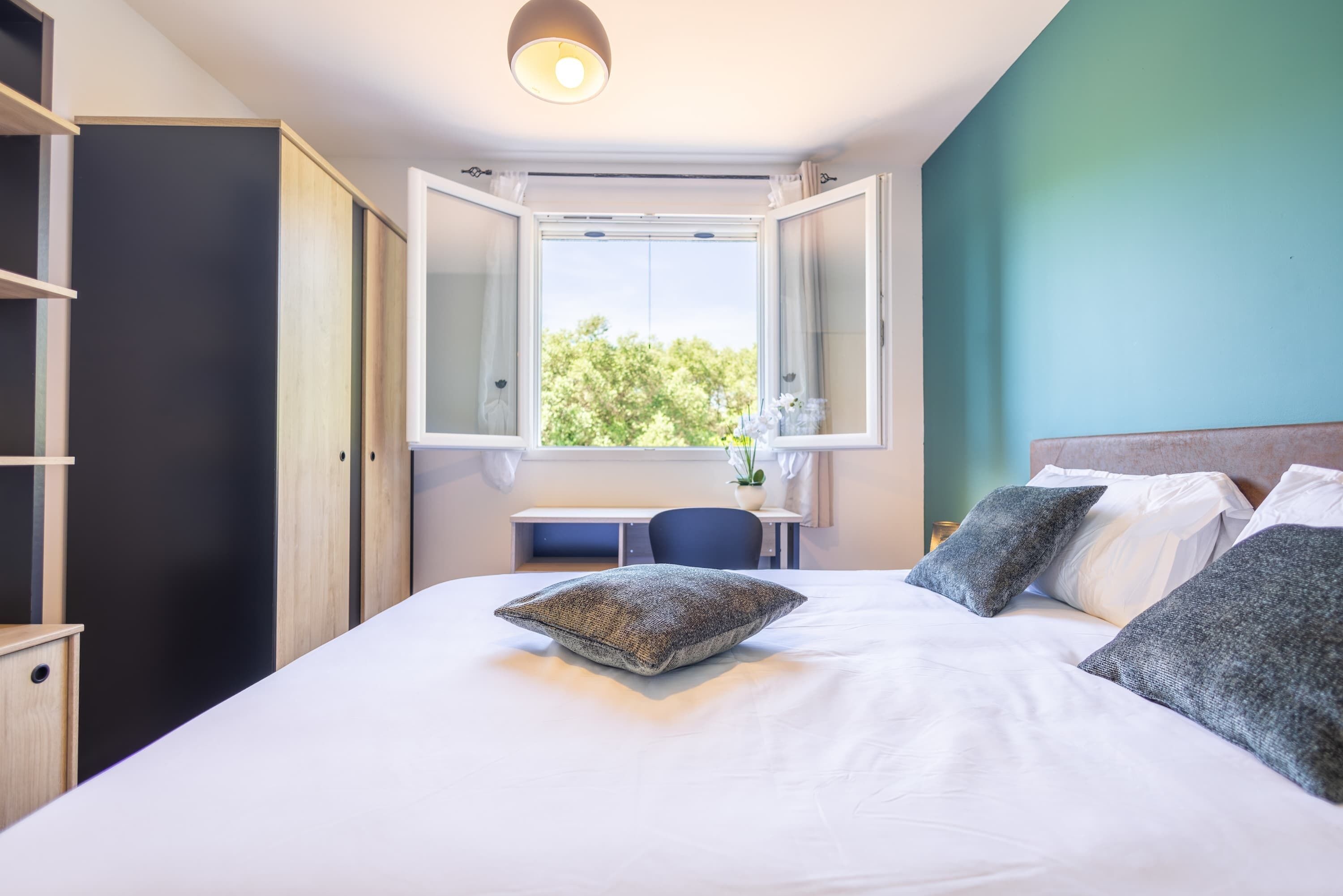Ein Schlafzimmer mit Bett, Kissen, Schreibtisch und Fenster.