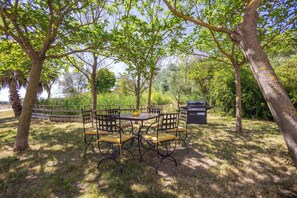 A table with chairs and a grill in a shaded outdoor area surrounded by trees. - Air-conditioned apartment with outdoor space — Domaine Les Castans, between Agde and Pézenas (Saint-Thibéry)