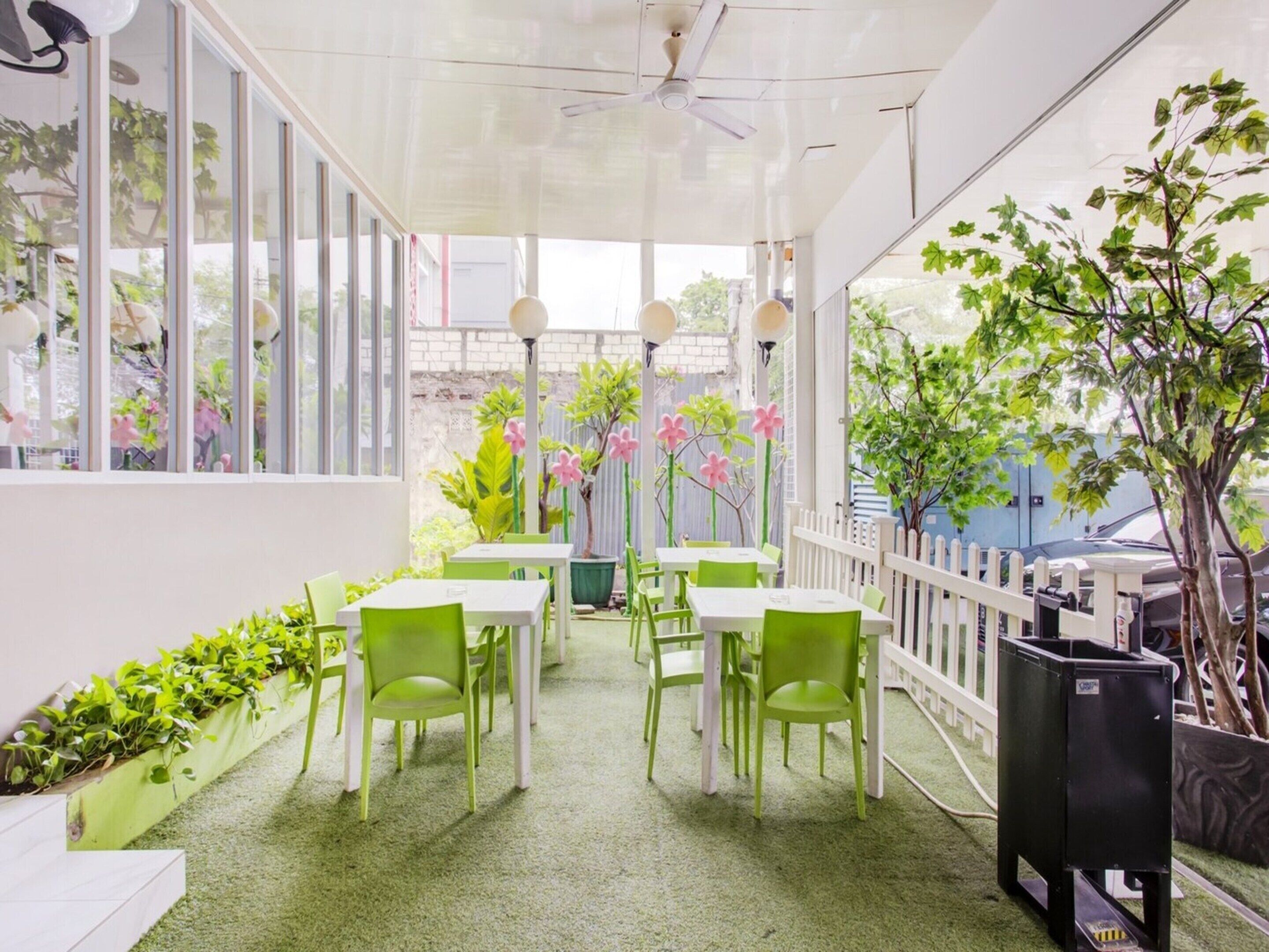 A covered outdoor seating area with green chairs and white tables, surrounded by potted plants and a white fence.