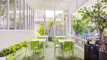 A covered outdoor seating area with green chairs and white tables, surrounded by potted plants and a white fence.
