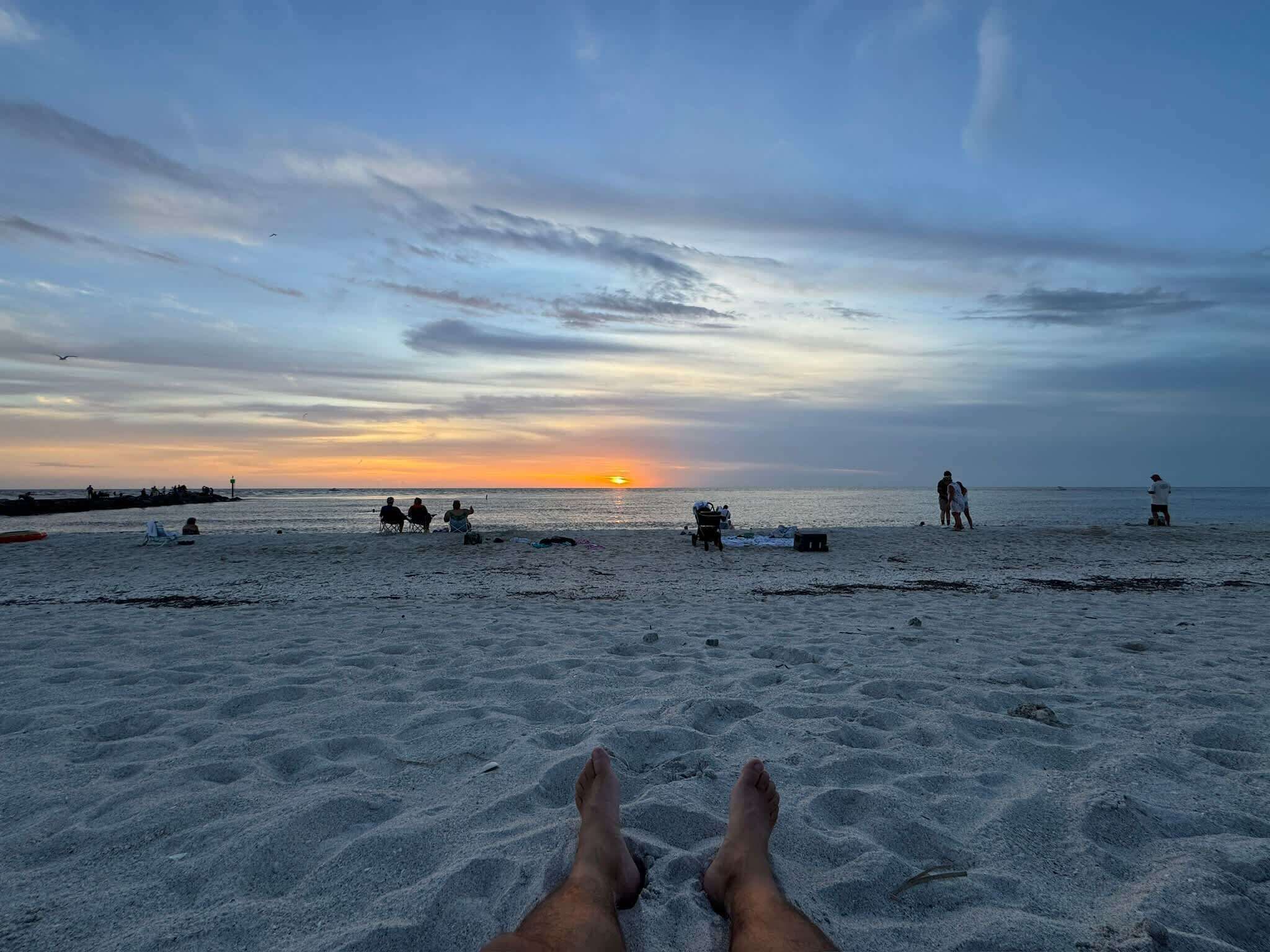 A beach with a sunset in the background, sandy ground, and a few people.