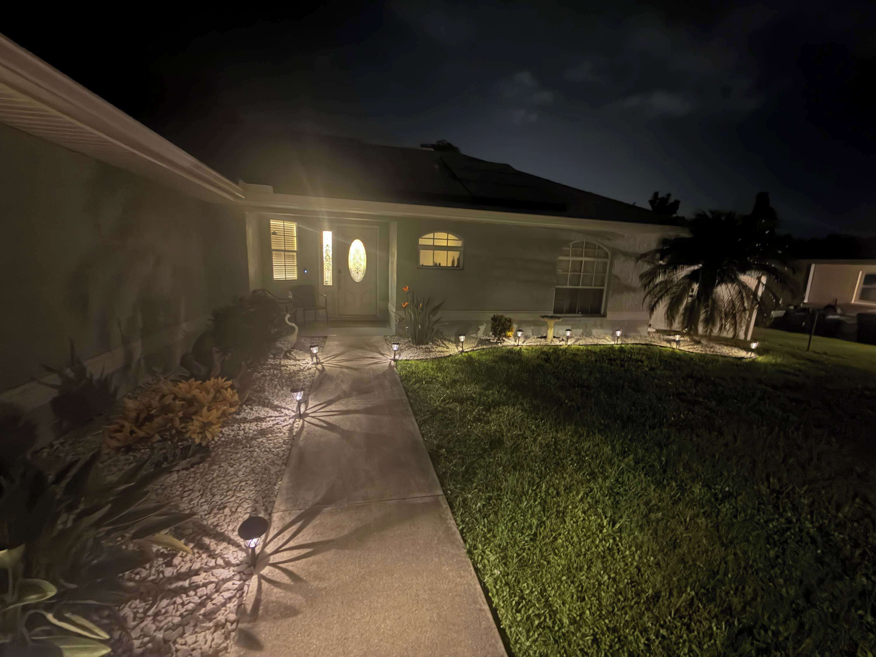 A house with a well-lit entrance, a paved driveway, and a landscaped front yard with a palm tree.