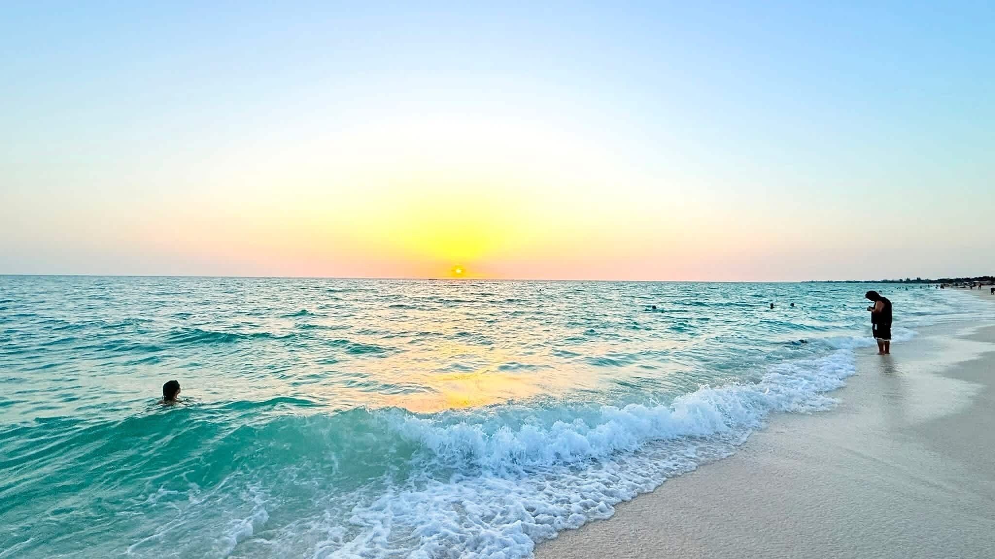 A beach with waves at sunset, a person standing on the sand, and another in the water.