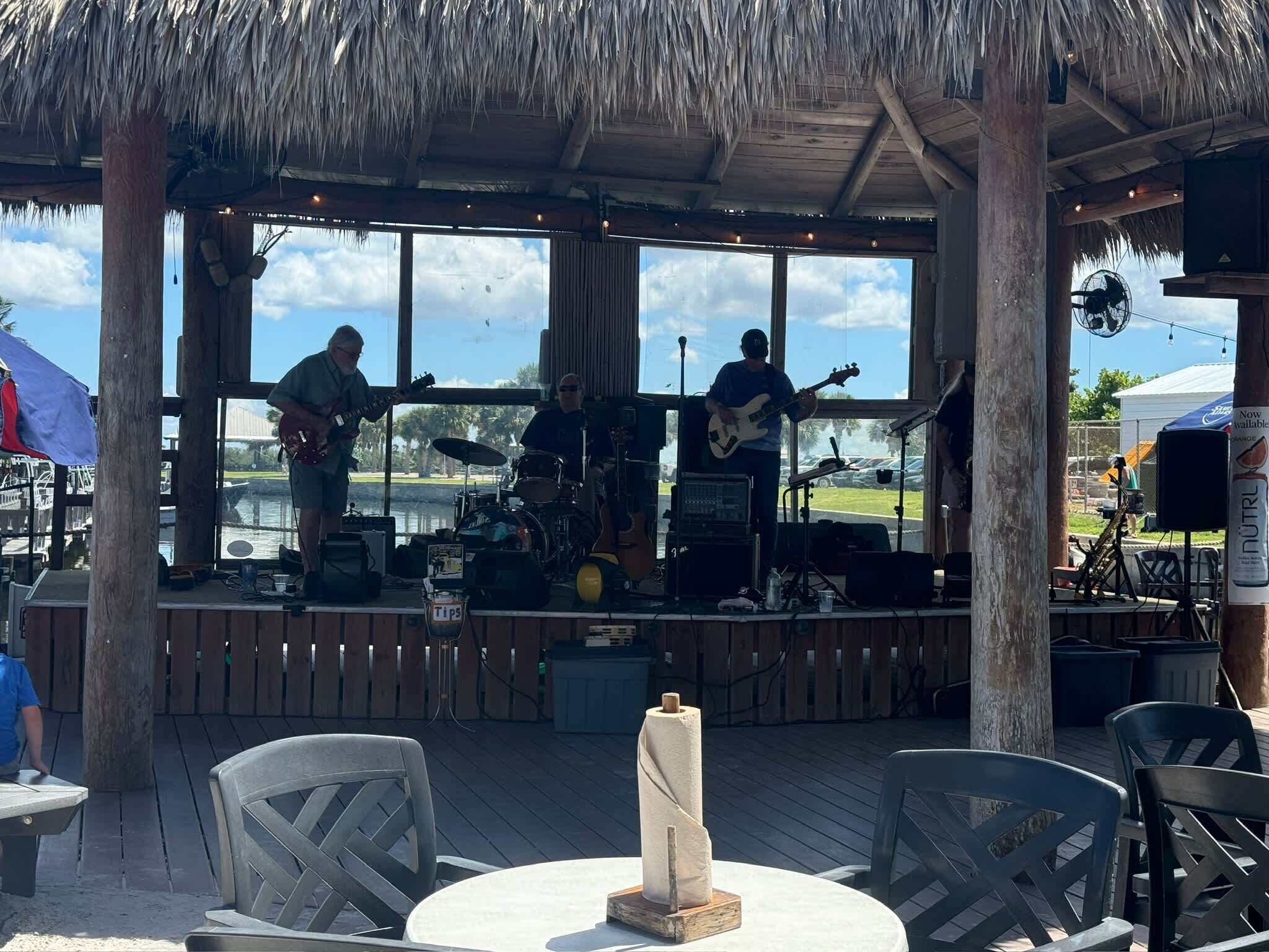 An outdoor music performance area with a thatched roof, a stage with musical instruments, and seating for an audience.