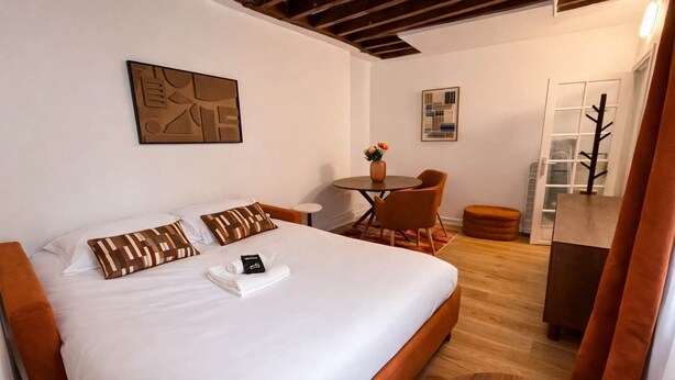 Une chambre d’hôtel avec un grand lit, une petite table ronde avec des chaises, une plante en pot et un banc en bois.