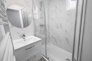 Ein modernes Badezimmer mit Duschkabine aus Glas, Wandverkleidung aus weißem Marmor, rundem Spiegel und Waschbecken mit einer Schublade.