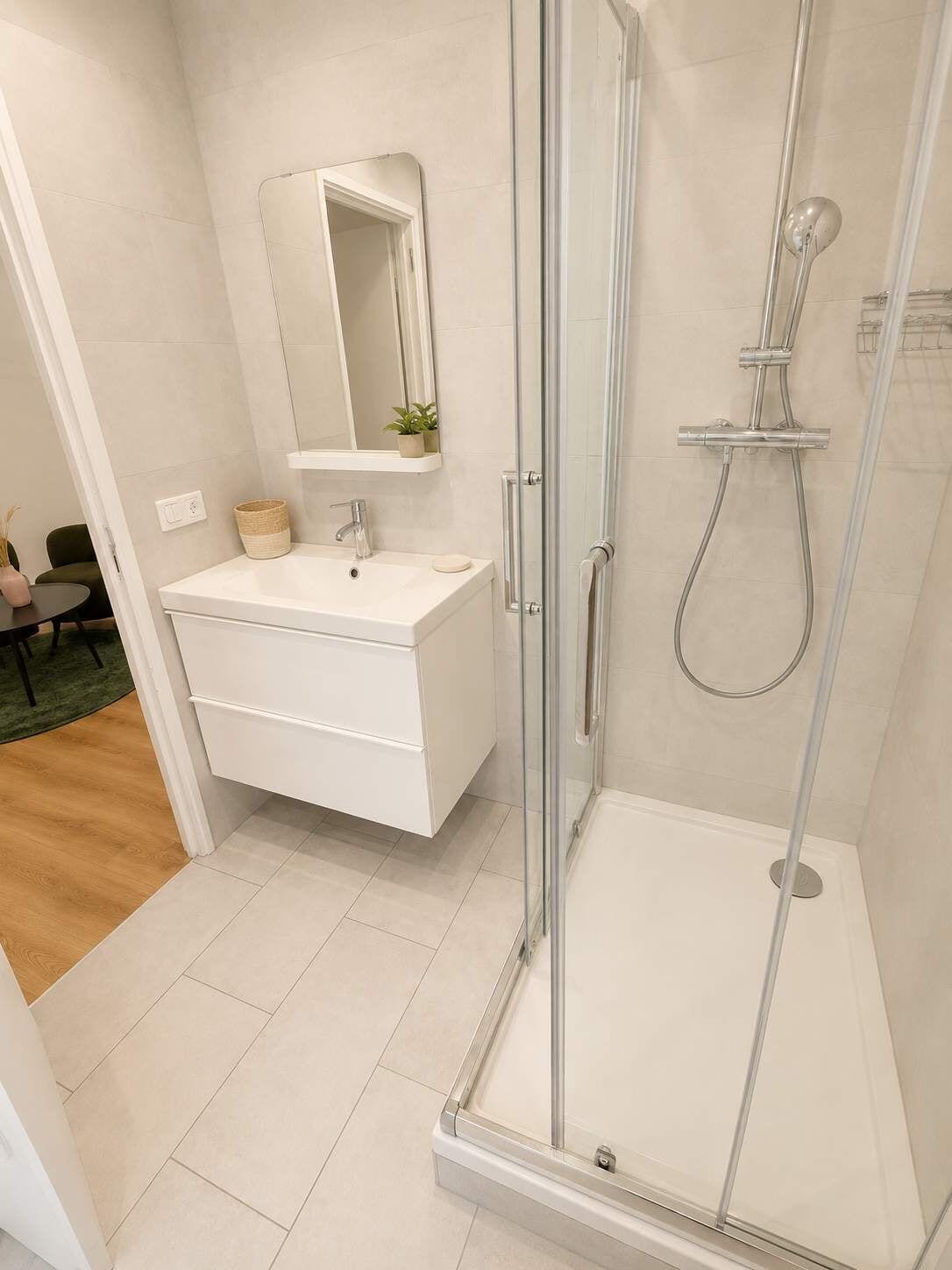 Une salle de bain moderne avec une douche à l’italienne, un meuble-lavabo blanc avec un miroir et une petite plante posée sur le meuble.