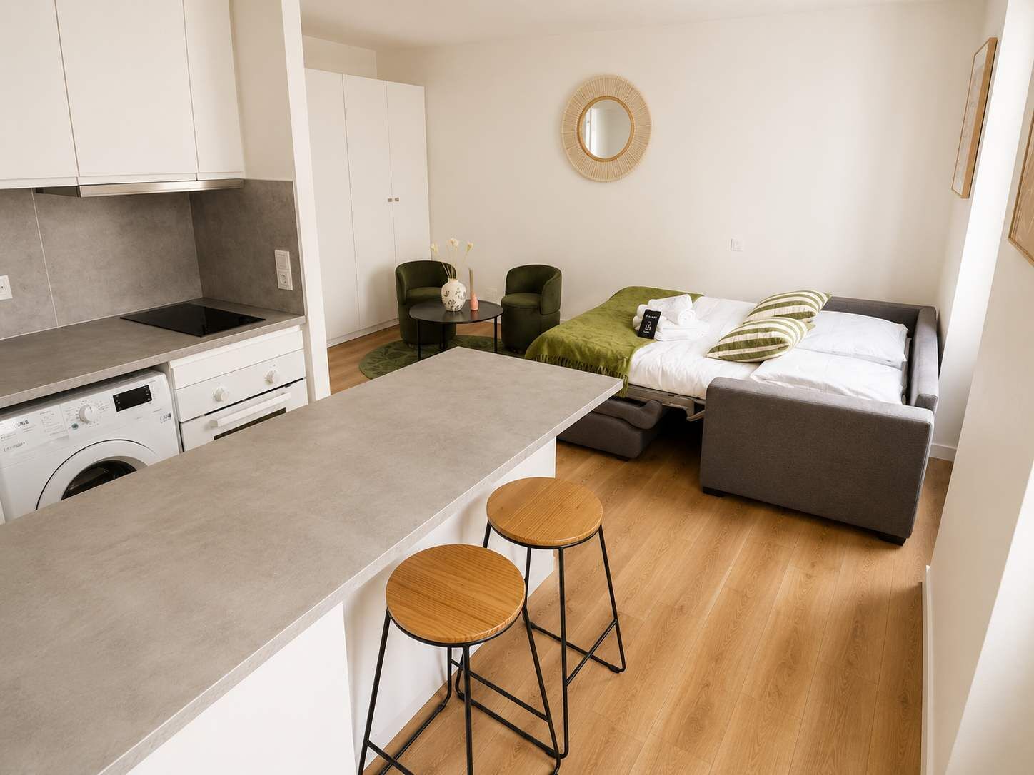 A compact living space with a kitchenette, a bed, a round mirror, and two stools.