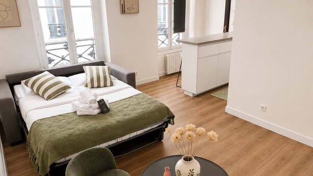 A modern hotel room with a bed, a sitting area with a round table, and a kitchenette in the background. - Charmant Studio Paris 11 - faubourg du temple (Paris)