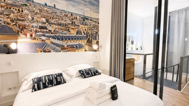 A hotel room with a bed, a painting of a cityscape, a balcony with a view, and a welcome sign. - Duplex Design Montparnasse - Baby-foot - 6P (Paris)