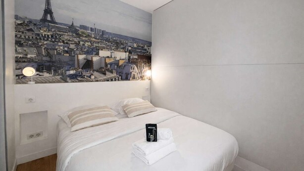 A hotel room with a bed, a painting of a cityscape, and a nightstand with a lamp. - Appartement Élégant -  Montparnasse - 6 Personnes (Paris)