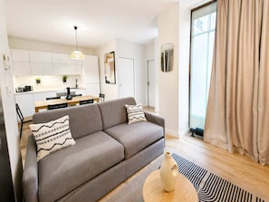 Ein modernes Wohnzimmer mit einem grauen Sofa, einem kleinen Tisch mit einer Vase und einer Küche im Hintergrund.