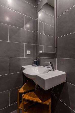 Ein modernes Badezimmer mit dunklen Fliesen, einem weißen Waschbecken, einem Spiegel und einem Holzstuhl.