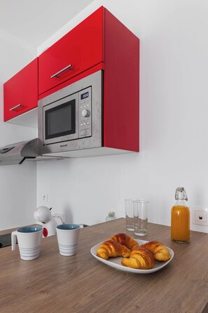 Eine Küche mit roten Schränken, einer Mikrowelle, einem Teller mit Croissants, zwei Tassen und einer Orangensaftflasche.