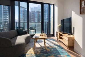 A modern living room with a sofa, a coffee table, a TV, and a large window with a city view. - Skyline Stays Southbank Rooftop Pool Apt (Southbank)