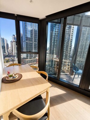 A modern dining area with a table and chairs, large windows with a city view, and a small balcony with a chair. - Skyline Stays Southbank Rooftop Pool Apt (Southbank)