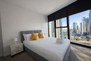 A bedroom with a large bed, two pillows, a nightstand, and a view of a cityscape through large windows. - Skyline Stays Southbank Rooftop Pool Apt (Southbank)
