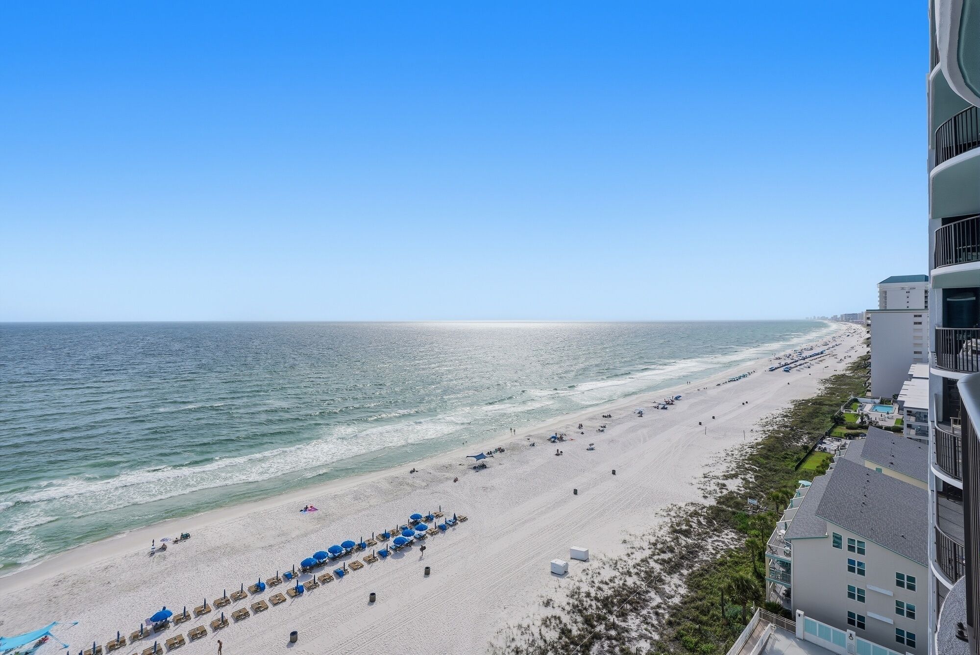A beach with rows of sun loungers and umbrellas, a clear sky, and a building with balconies overlooking the beach.