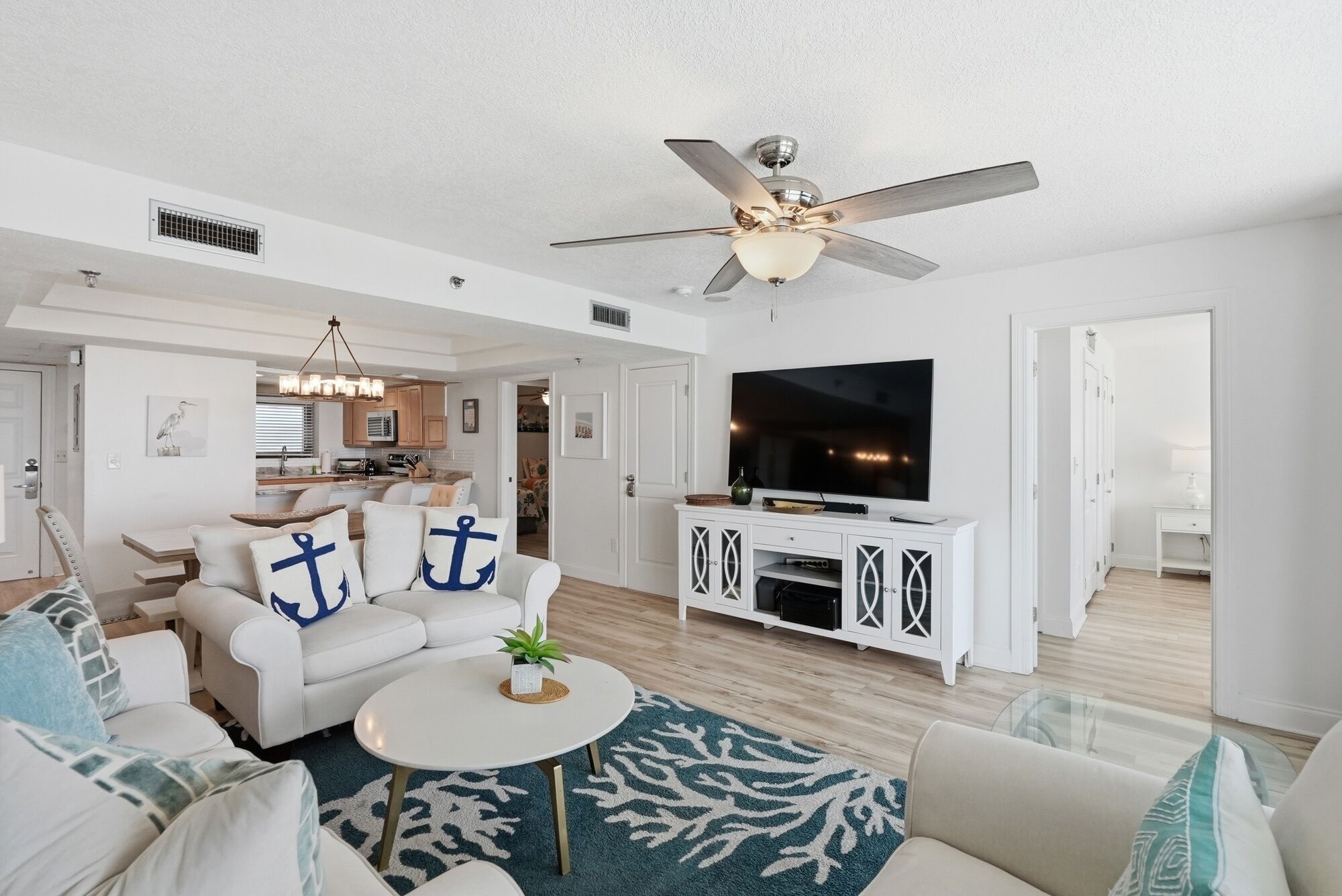 A spacious living room with a ceiling fan, a flat-screen TV, and a dining area with a kitchen in the background.