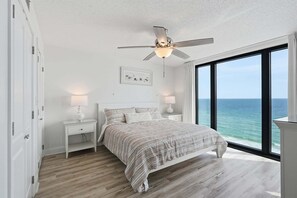 A bedroom with a bed, two bedside tables, a ceiling fan, a window with a view of the ocean, and a framed picture on the wall. - Watercrest 1404 - Free Beach Chairs! (Panama City)