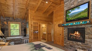 A log cabin interior with a stone fireplace, a television mounted above it, a large bed with a forest-themed bedspread, and a stone wall. - Crazy Horse (Sevierville)