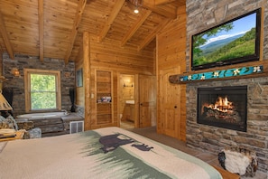 A log cabin interior with a stone fireplace, a television mounted above it, a large bed with a forest-themed bedspread, and a stone wall. - Crazy Horse (Sevierville)