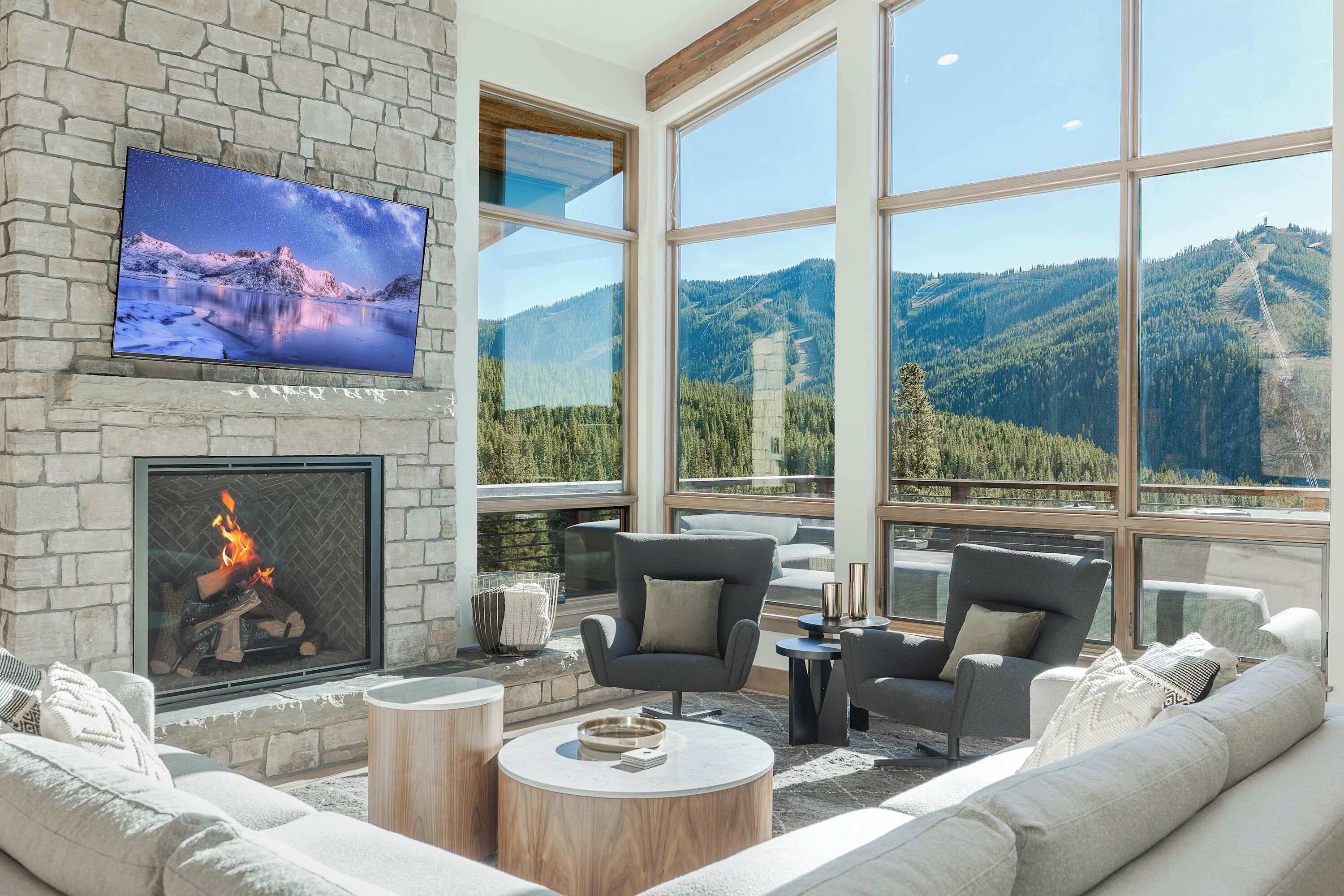A modern living room with a stone fireplace, comfortable seating, a flat-screen TV, and large windows offering mountain views.