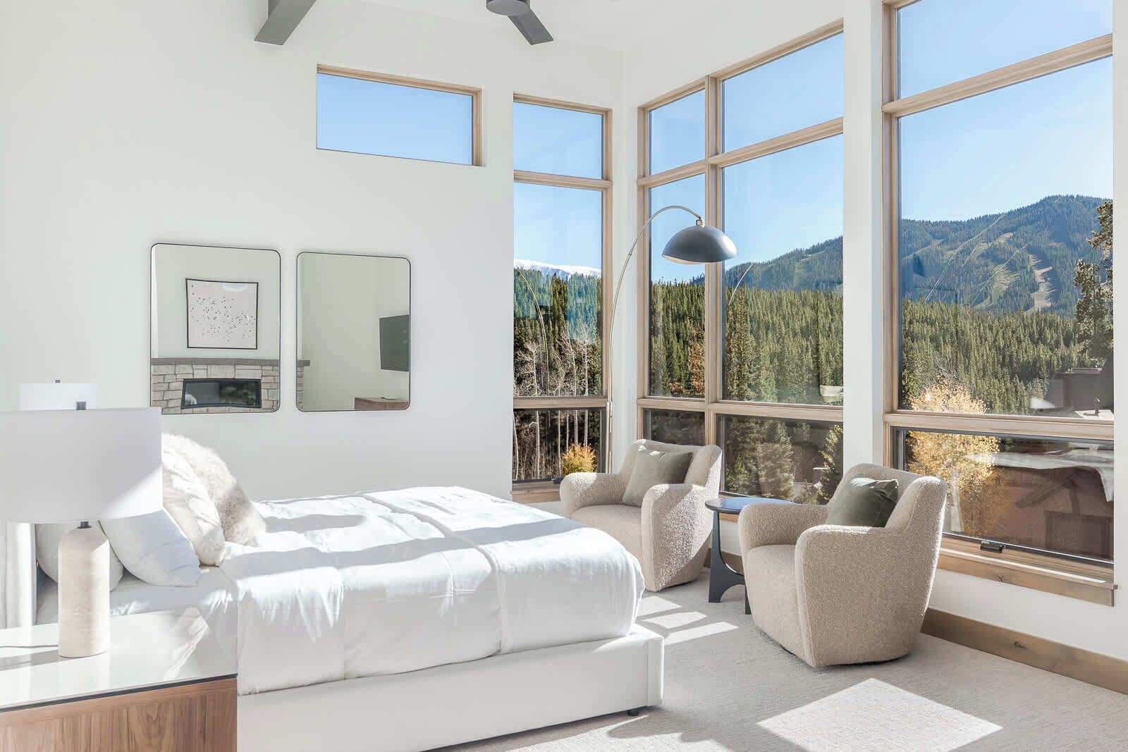 A bedroom with a bed, two chairs, a desk, a lamp, and large windows with a mountain view.